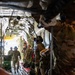 Military personnel in MultiCam/OCP camouflage uniforms are gathered inside the cargo hold of a large military transport aircraft, likely a C-130 Hercules. Several soldiers wearing communications headsets are visible, with the rear cargo ramp open, allowing natural light to illuminate the interior. The scene appears to depict a pre-mission briefing, loading, or aeromedical evacuation preparation.
