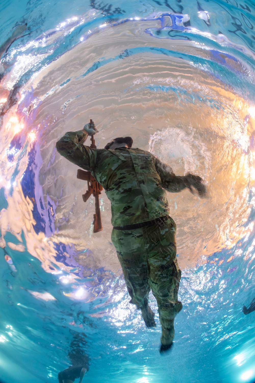 An underwater photograph taken from below the water surface looking up at a soldier in camouflage uniform swimming or treading water while holding a rifle above the waterline. The image is captured with a wide-angle or fisheye lens, creating a circular distortion effect with the water surface visible as a shimmering, reflective boundary. This appears to be a military water survival or combat swimming training exercise conducted in an indoor pool.