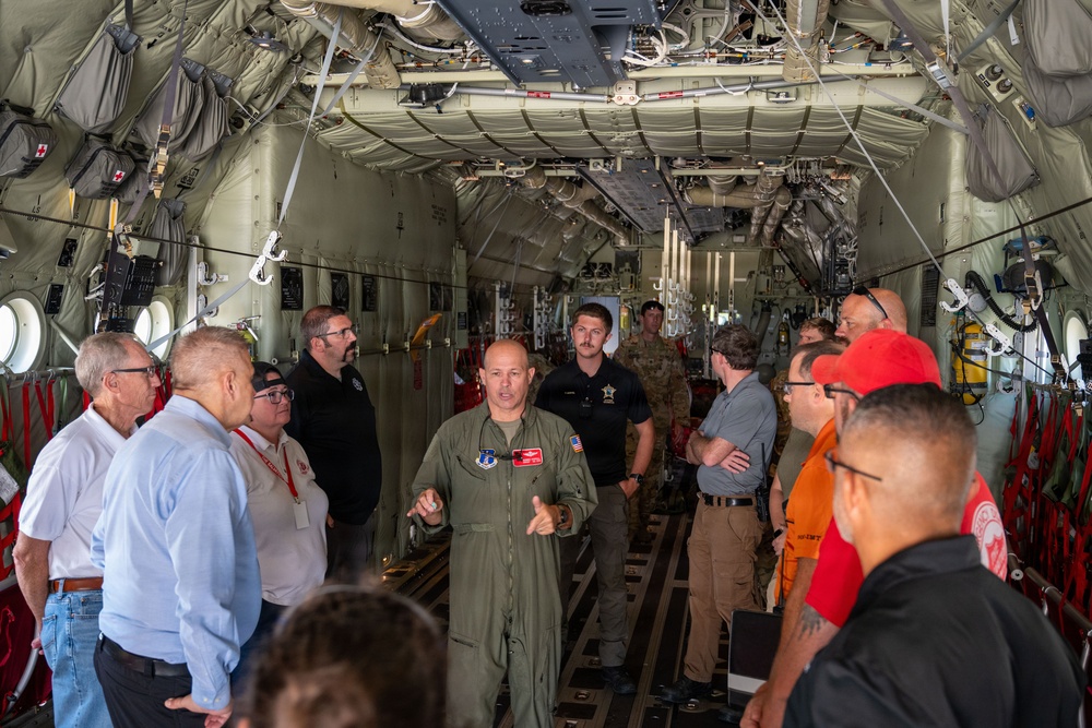 A military aviator in an olive green flight suit is briefing a group of approximately 15 civilians and personnel inside the cargo hold of a large military transport aircraft, likely a C-130 or similar aircraft. The speaker appears to be gesturing with both hands as he addresses the group gathered on both sides of the cargo bay. The setting suggests an official tour, orientation, or community outreach event.