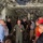 A military aviator in an olive green flight suit is briefing a group of approximately 15 civilians and personnel inside the cargo hold of a large military transport aircraft, likely a C-130 or similar aircraft. The speaker appears to be gesturing with both hands as he addresses the group gathered on both sides of the cargo bay. The setting suggests an official tour, orientation, or community outreach event.