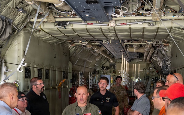 A military aviator in an olive green flight suit is briefing a group of approximately 15 civilians and personnel inside the cargo hold of a large military transport aircraft, likely a C-130 or similar aircraft. The speaker appears to be gesturing with both hands as he addresses the group gathered on both sides of the cargo bay. The setting suggests an official tour, orientation, or community outreach event.