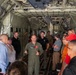 A military aviator in an olive green flight suit is briefing a group of approximately 15 civilians and personnel inside the cargo hold of a large military transport aircraft, likely a C-130 or similar aircraft. The speaker appears to be gesturing with both hands as he addresses the group gathered on both sides of the cargo bay. The setting suggests an official tour, orientation, or community outreach event.