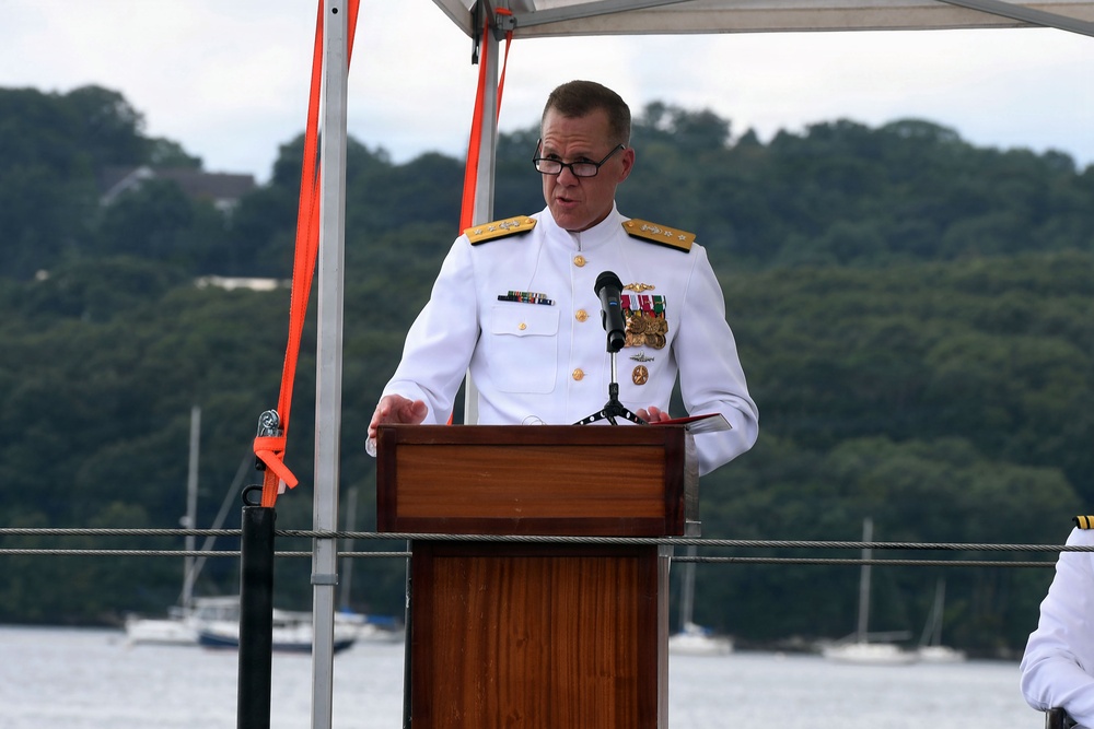 A senior naval officer in white dress uniform is speaking at a podium during what appears to be an official military ceremony. The event is taking place outdoors on or near a waterfront, with a body of water, sailboats, and tree-covered hills visible in the background. The officer displays multiple medals and insignia consistent with a high-ranking admiral.