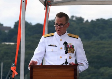 A senior naval officer in white dress uniform is speaking at a podium during what appears to be an official military ceremony. The event is taking place outdoors on or near a waterfront, with a body of water, sailboats, and tree-covered hills visible in the background. The officer displays multiple medals and insignia consistent with a high-ranking admiral.