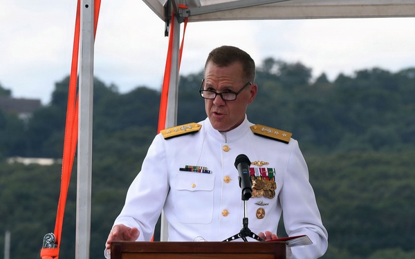 A senior naval officer in white dress uniform is speaking at a podium during what appears to be an official military ceremony. The event is taking place outdoors on or near a waterfront, with a body of water, sailboats, and tree-covered hills visible in the background. The officer displays multiple medals and insignia consistent with a high-ranking admiral.