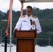 A senior naval officer in white dress uniform is speaking at a podium during what appears to be an official military ceremony. The event is taking place outdoors on or near a waterfront, with a body of water, sailboats, and tree-covered hills visible in the background. The officer displays multiple medals and insignia consistent with a high-ranking admiral.