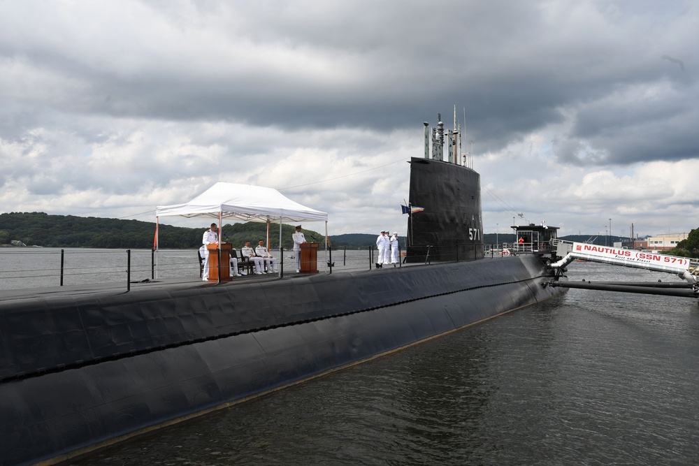 A formal military ceremony is taking place on the deck of the USS Nautilus (SSN 571), the world's first nuclear-powered submarine, docked at what appears to be the Submarine Force Museum in Groton, Connecticut. Naval personnel in white dress uniforms are gathered around podiums under a white canopy tent on the submarine's deck. The vessel's hull number '571' is visible on the sail, and a sign reading 'NAUTILUS (SSN 571)' is visible on the dock.