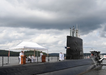 A formal military ceremony is taking place on the deck of the USS Nautilus (SSN 571), the world's first nuclear-powered submarine, docked at what appears to be the Submarine Force Museum in Groton, Connecticut. Naval personnel in white dress uniforms are gathered around podiums under a white canopy tent on the submarine's deck. The vessel's hull number '571' is visible on the sail, and a sign reading 'NAUTILUS (SSN 571)' is visible on the dock.