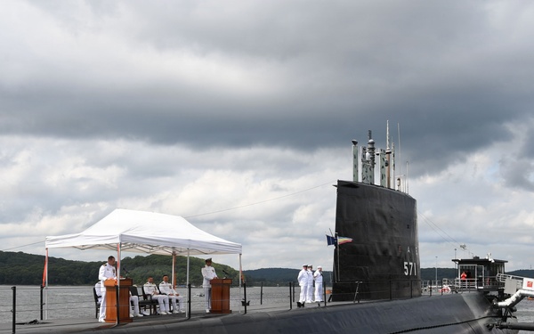 A formal military ceremony is taking place on the deck of the USS Nautilus (SSN 571), the world's first nuclear-powered submarine, docked at what appears to be the Submarine Force Museum in Groton, Connecticut. Naval personnel in white dress uniforms are gathered around podiums under a white canopy tent on the submarine's deck. The vessel's hull number '571' is visible on the sail, and a sign reading 'NAUTILUS (SSN 571)' is visible on the dock.