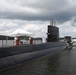 A formal military ceremony is taking place on the deck of the USS Nautilus (SSN 571), the world's first nuclear-powered submarine, docked at what appears to be the Submarine Force Museum in Groton, Connecticut. Naval personnel in white dress uniforms are gathered around podiums under a white canopy tent on the submarine's deck. The vessel's hull number '571' is visible on the sail, and a sign reading 'NAUTILUS (SSN 571)' is visible on the dock.