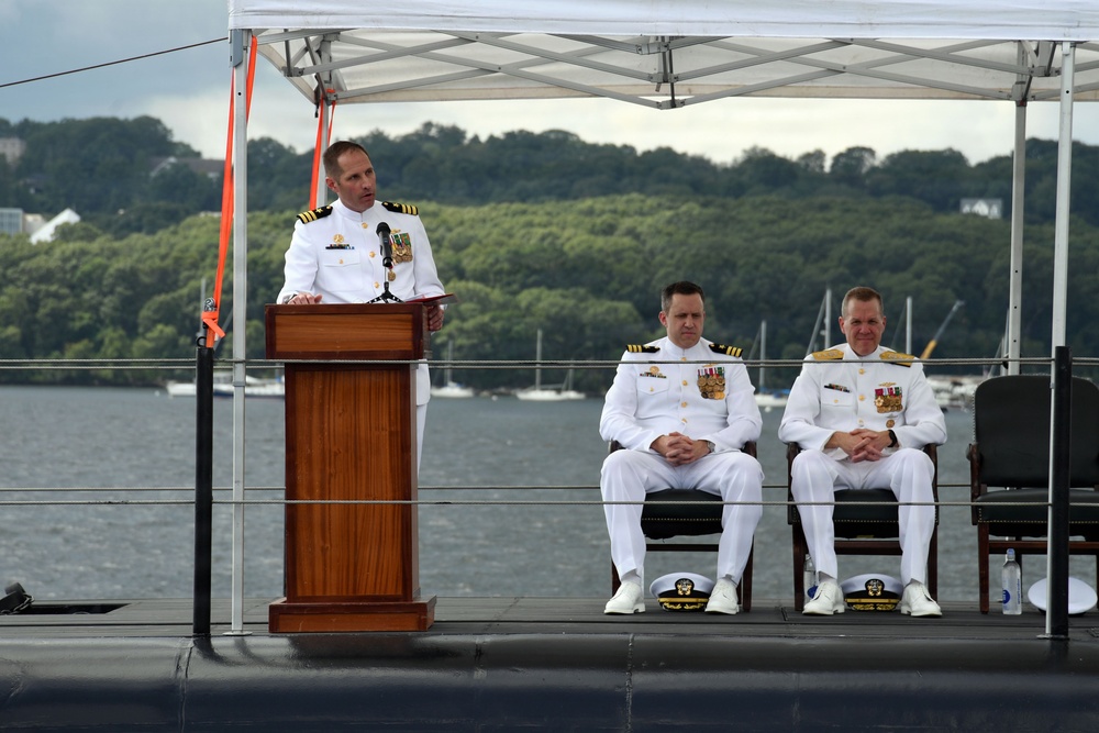 Three U.S. Navy officers in white dress uniforms are participating in a formal military ceremony, likely a change of command or commissioning event, conducted outdoors on what appears to be the deck of a submarine. One officer stands at a wooden podium speaking, while two others are seated behind him. The ceremony is taking place near a waterway with sailboats and a tree-lined shore visible in the background.