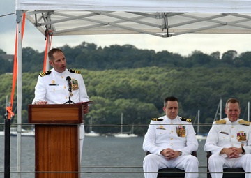 Three U.S. Navy officers in white dress uniforms are participating in a formal military ceremony, likely a change of command or commissioning event, conducted outdoors on what appears to be the deck of a submarine. One officer stands at a wooden podium speaking, while two others are seated behind him. The ceremony is taking place near a waterway with sailboats and a tree-lined shore visible in the background.