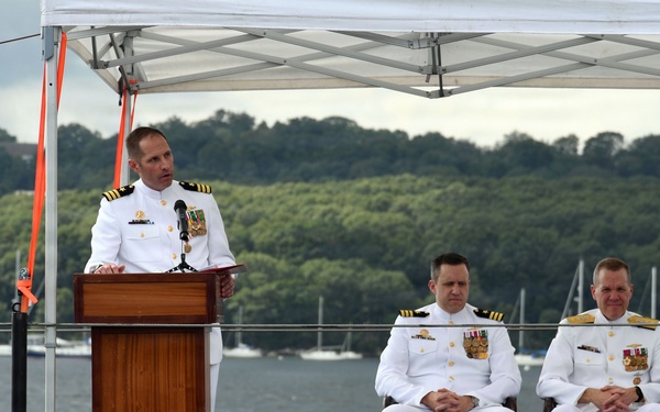 Three U.S. Navy officers in white dress uniforms are participating in a formal military ceremony, likely a change of command or commissioning event, conducted outdoors on what appears to be the deck of a submarine. One officer stands at a wooden podium speaking, while two others are seated behind him. The ceremony is taking place near a waterway with sailboats and a tree-lined shore visible in the background.