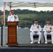 Three U.S. Navy officers in white dress uniforms are participating in a formal military ceremony, likely a change of command or commissioning event, conducted outdoors on what appears to be the deck of a submarine. One officer stands at a wooden podium speaking, while two others are seated behind him. The ceremony is taking place near a waterway with sailboats and a tree-lined shore visible in the background.