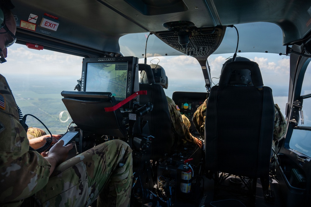 Interior view of a military helicopter in flight, showing crew members in multicam/OCP uniforms operating the aircraft. At least two personnel are visible: one in the left rear area reviewing a smartphone, and at least one pilot/crew member in the forward cockpit area. The aircraft is flying over a green, partially forested landscape with scattered clouds visible through the windscreen.