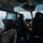 Interior view of a military helicopter in flight, showing crew members in multicam/OCP uniforms operating the aircraft. At least two personnel are visible: one in the left rear area reviewing a smartphone, and at least one pilot/crew member in the forward cockpit area. The aircraft is flying over a green, partially forested landscape with scattered clouds visible through the windscreen.