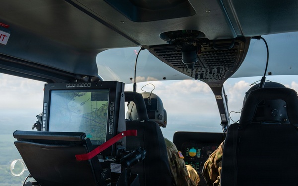 Interior view of a military helicopter in flight, showing crew members in multicam/OCP uniforms operating the aircraft. At least two personnel are visible: one in the left rear area reviewing a smartphone, and at least one pilot/crew member in the forward cockpit area. The aircraft is flying over a green, partially forested landscape with scattered clouds visible through the windscreen.