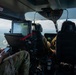 Interior view of a military helicopter in flight, showing crew members in multicam/OCP uniforms operating the aircraft. At least two personnel are visible: one in the left rear area reviewing a smartphone, and at least one pilot/crew member in the forward cockpit area. The aircraft is flying over a green, partially forested landscape with scattered clouds visible through the windscreen.