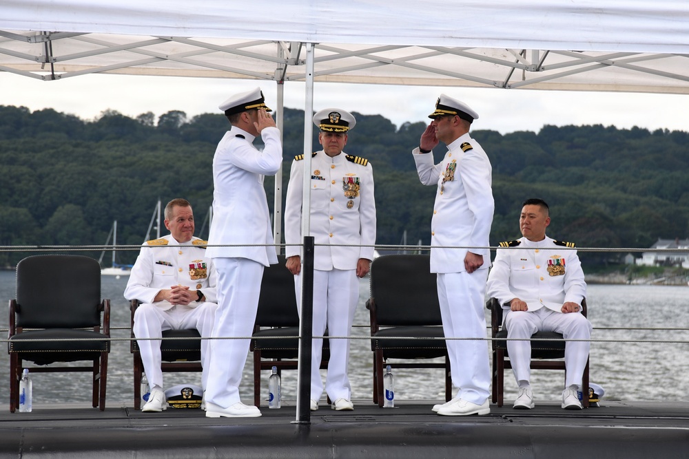 A formal U.S. Navy military ceremony, likely a change of command, taking place on the deck of a submarine at a waterfront location. Five naval officers in white dress uniforms are present, with two officers exchanging salutes in the center foreground while two others are seated and one stands at attention in the background. The setting includes a white canopy tent, dark submarine deck surface, and a tree-lined waterway in the background.