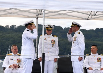 A formal U.S. Navy military ceremony, likely a change of command, taking place on the deck of a submarine at a waterfront location. Five naval officers in white dress uniforms are present, with two officers exchanging salutes in the center foreground while two others are seated and one stands at attention in the background. The setting includes a white canopy tent, dark submarine deck surface, and a tree-lined waterway in the background.