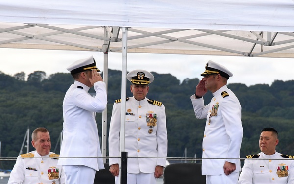 A formal U.S. Navy military ceremony, likely a change of command, taking place on the deck of a submarine at a waterfront location. Five naval officers in white dress uniforms are present, with two officers exchanging salutes in the center foreground while two others are seated and one stands at attention in the background. The setting includes a white canopy tent, dark submarine deck surface, and a tree-lined waterway in the background.