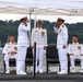A formal U.S. Navy military ceremony, likely a change of command, taking place on the deck of a submarine at a waterfront location. Five naval officers in white dress uniforms are present, with two officers exchanging salutes in the center foreground while two others are seated and one stands at attention in the background. The setting includes a white canopy tent, dark submarine deck surface, and a tree-lined waterway in the background.