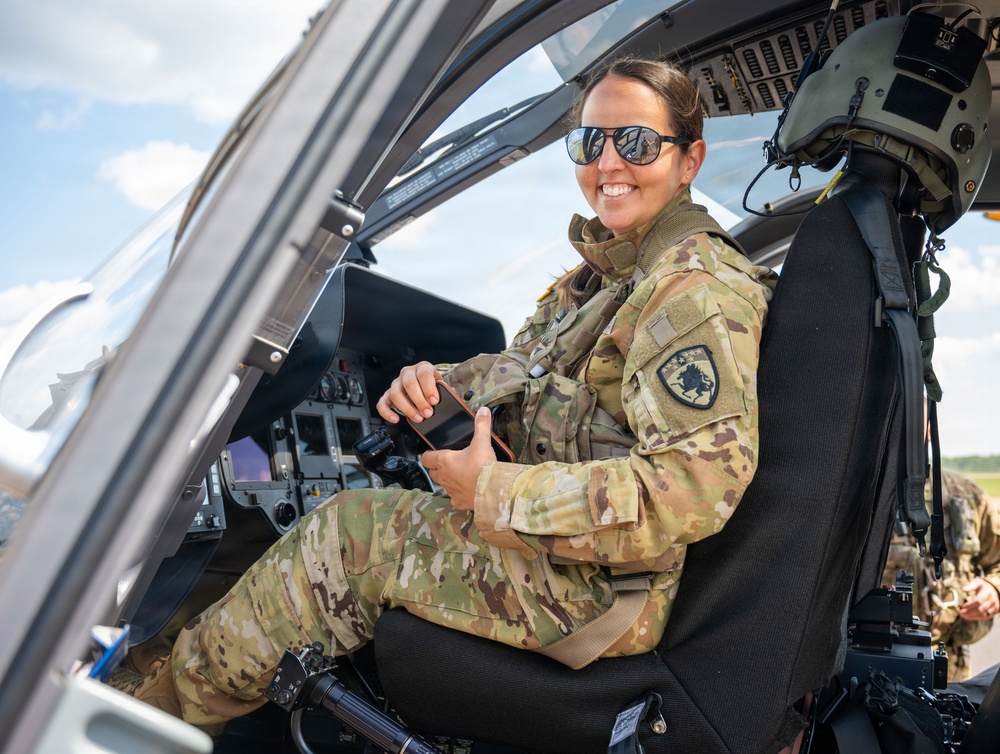A female military service member in OCP (Operational Camouflage Pattern) uniform is seated in the cockpit of a military helicopter, smiling at the camera. She is wearing aviator-style sunglasses and has a unit patch visible on her left shoulder. The setting appears to be an airfield on a partly cloudy day.
