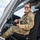 A female military service member in OCP (Operational Camouflage Pattern) uniform is seated in the cockpit of a military helicopter, smiling at the camera. She is wearing aviator-style sunglasses and has a unit patch visible on her left shoulder. The setting appears to be an airfield on a partly cloudy day.