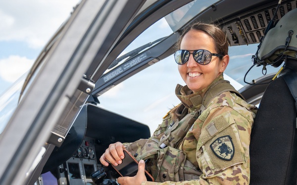 A female military service member in OCP (Operational Camouflage Pattern) uniform is seated in the cockpit of a military helicopter, smiling at the camera. She is wearing aviator-style sunglasses and has a unit patch visible on her left shoulder. The setting appears to be an airfield on a partly cloudy day.