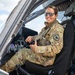 A female military service member in OCP (Operational Camouflage Pattern) uniform is seated in the cockpit of a military helicopter, smiling at the camera. She is wearing aviator-style sunglasses and has a unit patch visible on her left shoulder. The setting appears to be an airfield on a partly cloudy day.