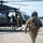A U.S. Army soldier in full flight gear walks away from a military helicopter on a tarmac, carrying a flight helmet in hand. Additional soldiers in camouflage uniforms are visible boarding or working near the open side door of the dark-colored helicopter. The scene appears to depict pre-flight or post-flight operations at a military airfield.