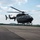 A dark gray/black military-style helicopter is captured in a low hover or just lifting off from a paved tarmac surface. The main rotor blades are visibly spinning, shown by motion blur. The setting appears to be a military or civilian airfield surrounded by green treelines and rolling hills under a partly cloudy sky.
