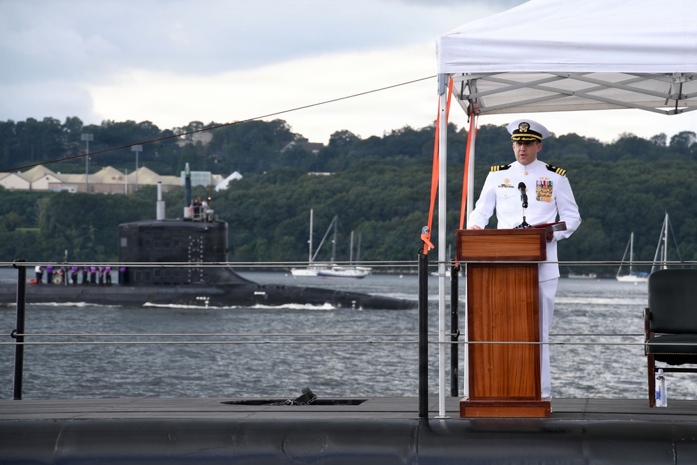 A U.S. Navy officer in dress white uniform stands at a wooden podium delivering a speech at what appears to be an official military ceremony. The event is taking place on the deck of a submarine, with another submarine visible transiting the waterway in the background. A white canopy tent provides partial cover over the speaking area.