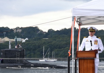 A U.S. Navy officer in dress white uniform stands at a wooden podium delivering a speech at what appears to be an official military ceremony. The event is taking place on the deck of a submarine, with another submarine visible transiting the waterway in the background. A white canopy tent provides partial cover over the speaking area.