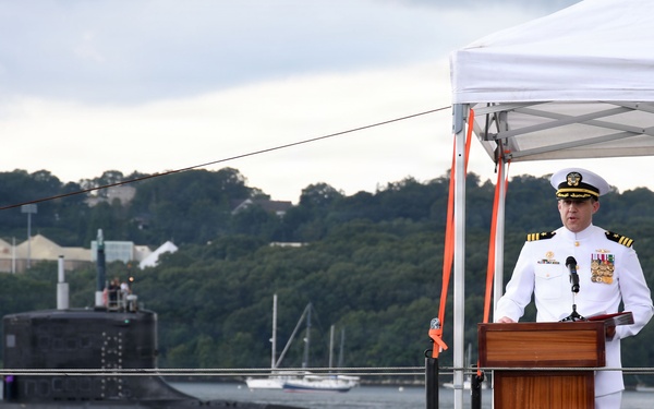 A U.S. Navy officer in dress white uniform stands at a wooden podium delivering a speech at what appears to be an official military ceremony. The event is taking place on the deck of a submarine, with another submarine visible transiting the waterway in the background. A white canopy tent provides partial cover over the speaking area.