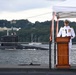 A U.S. Navy officer in dress white uniform stands at a wooden podium delivering a speech at what appears to be an official military ceremony. The event is taking place on the deck of a submarine, with another submarine visible transiting the waterway in the background. A white canopy tent provides partial cover over the speaking area.