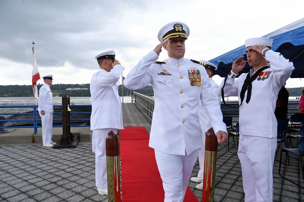 A formal U.S. Navy ceremony is taking place on a waterfront pier, with multiple naval personnel in dress white uniforms rendering salutes. A senior officer wearing a combination cover with gold insignia and numerous medals and ribbons is prominently centered, walking along a red carpet. The setting appears to be a change of command or similar official military ceremony.