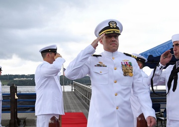 A formal U.S. Navy ceremony is taking place on a waterfront pier, with multiple naval personnel in dress white uniforms rendering salutes. A senior officer wearing a combination cover with gold insignia and numerous medals and ribbons is prominently centered, walking along a red carpet. The setting appears to be a change of command or similar official military ceremony.