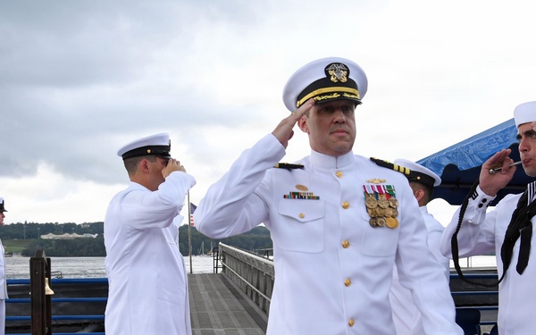 A formal U.S. Navy ceremony is taking place on a waterfront pier, with multiple naval personnel in dress white uniforms rendering salutes. A senior officer wearing a combination cover with gold insignia and numerous medals and ribbons is prominently centered, walking along a red carpet. The setting appears to be a change of command or similar official military ceremony.