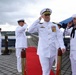 A formal U.S. Navy ceremony is taking place on a waterfront pier, with multiple naval personnel in dress white uniforms rendering salutes. A senior officer wearing a combination cover with gold insignia and numerous medals and ribbons is prominently centered, walking along a red carpet. The setting appears to be a change of command or similar official military ceremony.