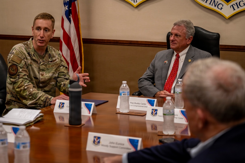 A U.S. Air Force officer in camouflage uniform is speaking at a formal briefing or meeting at what appears to be a military installation. A civilian in a gray suit with a red tie, identified by a nameplate as 'Rep. Buddy Carter, 1st Congressional District,' is seated across the table listening. The setting includes Air Force insignia, an American flag, and multiple nameplates indicating an official congressional visit.