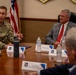 A U.S. Air Force officer in camouflage uniform is speaking at a formal briefing or meeting at what appears to be a military installation. A civilian in a gray suit with a red tie, identified by a nameplate as 'Rep. Buddy Carter, 1st Congressional District,' is seated across the table listening. The setting includes Air Force insignia, an American flag, and multiple nameplates indicating an official congressional visit.