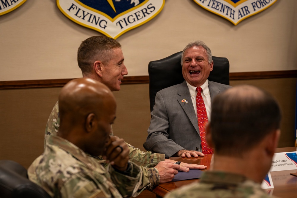 A meeting is taking place in what appears to be a military conference room, with military personnel in camouflage uniforms and a civilian in a gray suit sharing a moment of laughter. The setting is identified by insignia on the wall as a Flying Tigers / Fourteenth Air Force facility. A name placard partially visible reads 'Buddy Carter, Congressional District,' suggesting a congressional visit to a military installation.