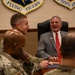 A meeting is taking place in what appears to be a military conference room, with military personnel in camouflage uniforms and a civilian in a gray suit sharing a moment of laughter. The setting is identified by insignia on the wall as a Flying Tigers / Fourteenth Air Force facility. A name placard partially visible reads 'Buddy Carter, Congressional District,' suggesting a congressional visit to a military installation.