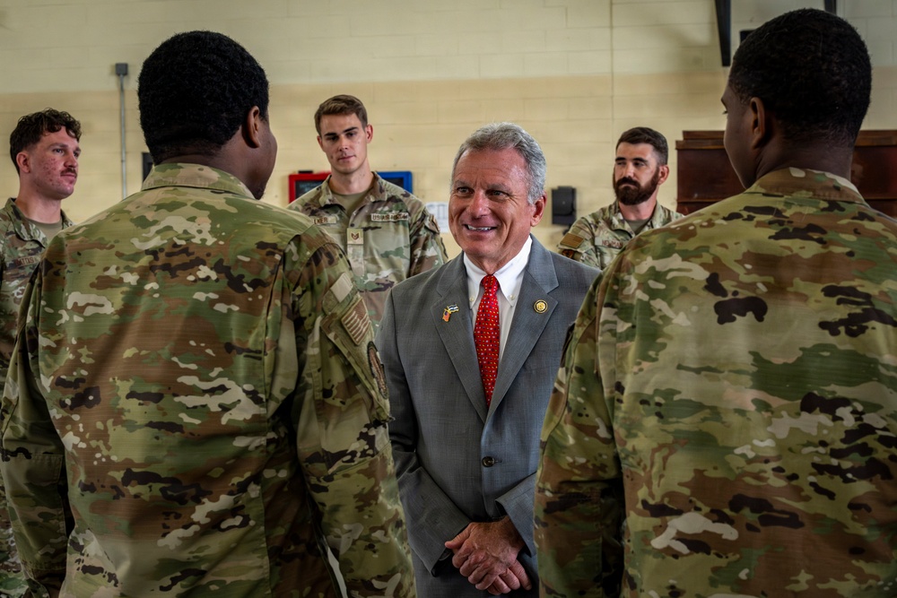 A civilian man in a gray suit with a red tie and lapel pin is engaged in conversation with several military personnel wearing OCP (Operational Camouflage Pattern) uniforms. The setting appears to be an indoor military facility or gymnasium. The interaction appears cordial and informal, suggesting a visit or meet-and-greet event.