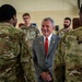 A civilian man in a gray suit with a red tie and lapel pin is engaged in conversation with several military personnel wearing OCP (Operational Camouflage Pattern) uniforms. The setting appears to be an indoor military facility or gymnasium. The interaction appears cordial and informal, suggesting a visit or meet-and-greet event.