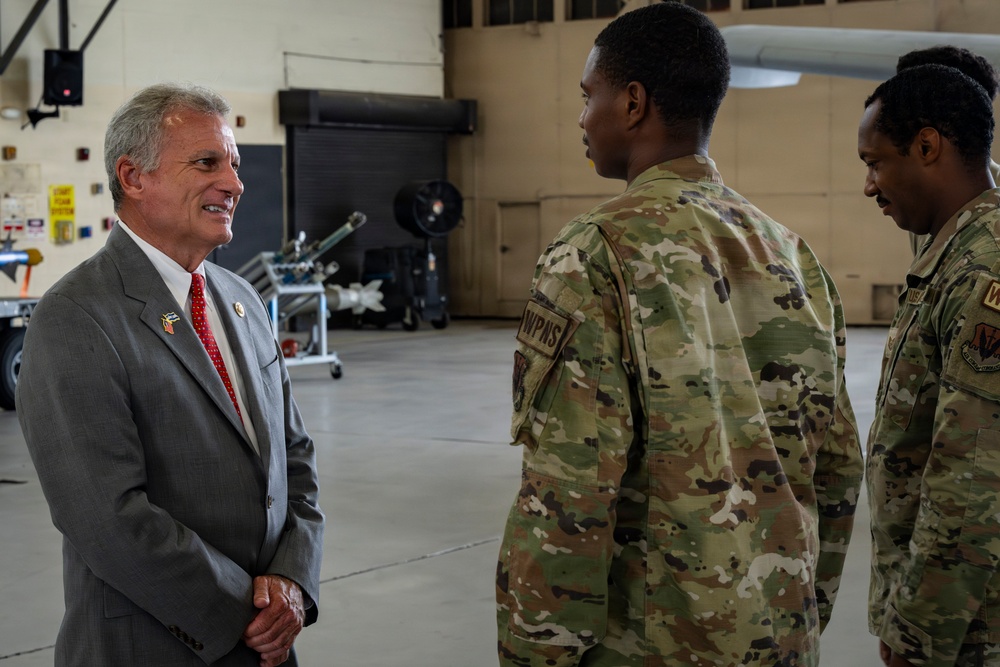 A civilian in a gray suit and red tie is engaged in conversation with two military personnel wearing OCP (Operational Camouflage Pattern) uniforms inside what appears to be a military aircraft hangar. The civilian appears to be a dignitary or official, identifiable by a lapel pin on his jacket. The setting includes weapons or ordnance equipment visible in the background.