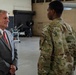 A civilian in a gray suit and red tie is engaged in conversation with two military personnel wearing OCP (Operational Camouflage Pattern) uniforms inside what appears to be a military aircraft hangar. The civilian appears to be a dignitary or official, identifiable by a lapel pin on his jacket. The setting includes weapons or ordnance equipment visible in the background.