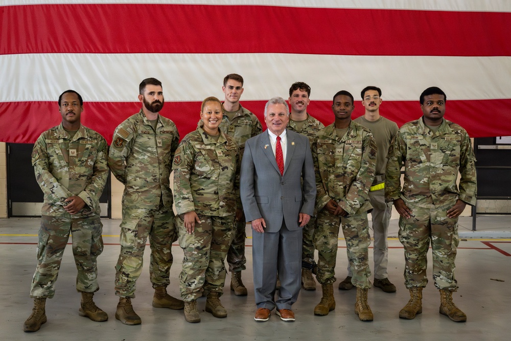A group of eight military personnel in OCP (Operational Camouflage Pattern) uniforms pose with one civilian man in a gray suit and red tie in front of a large American flag backdrop inside what appears to be a military hangar. The civilian is positioned in the center front of the group, suggesting a dignitary or official visit. The setting and attire indicate a formal commemorative or meet-and-greet occasion.