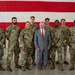 A group of eight military personnel in OCP (Operational Camouflage Pattern) uniforms pose with one civilian man in a gray suit and red tie in front of a large American flag backdrop inside what appears to be a military hangar. The civilian is positioned in the center front of the group, suggesting a dignitary or official visit. The setting and attire indicate a formal commemorative or meet-and-greet occasion.